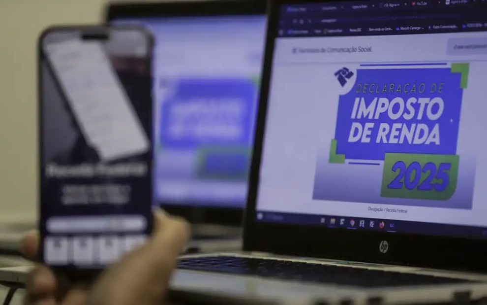 Notebook com a tela escrita "Imposto de Renda 2025" e um celular posicionado à frente.