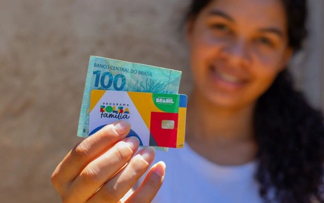Mulher segurando o cartão do Bolsa Família e uma cédula de cem reais