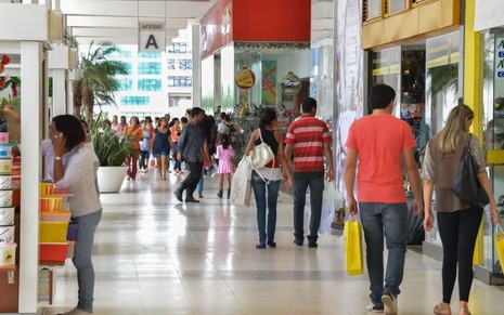 Pessoas caminham pelos corredores do shopping, carregando sacolas de compras nas mãos.